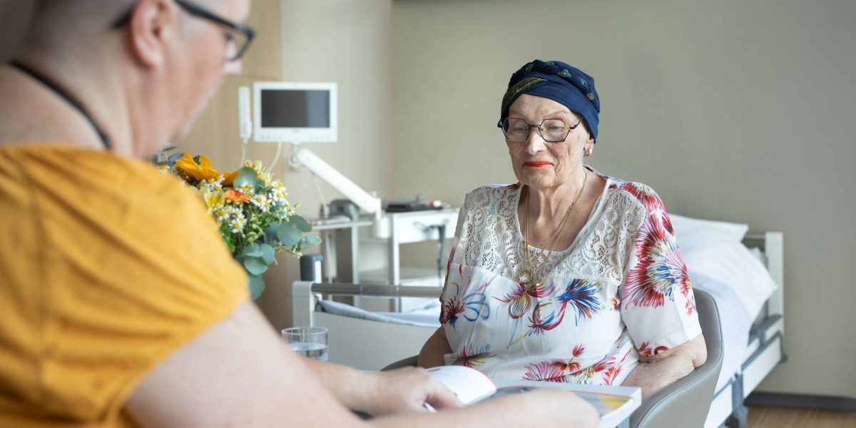 Ältere Frau mit Kopftuch sitzt an einem Tisch in einem Krankenhauszimmer, während eine andere Person ihr ein Buch zeigt.