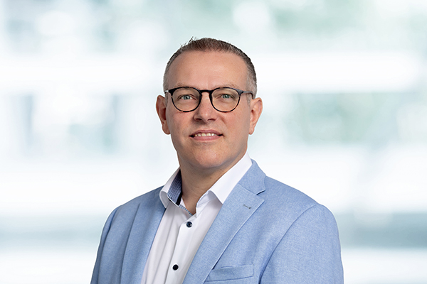 Ein Mann mit Brille in hellblauem Sakko lächelt vor unscharfem Hintergrund in die Kamera.