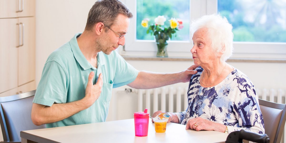 Pfleger spricht beruhigend mit älterer Frau am Tisch. Im Hintergrund Blumen, davor Becher und Pudding.