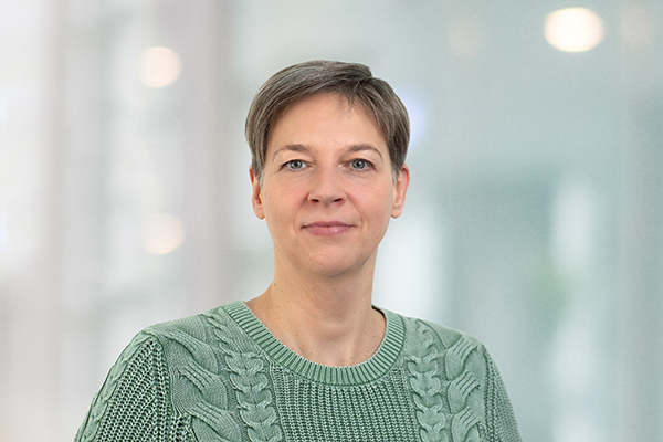 Porträt einer Person mit kurzem Haar in einem grünen Strickpullover vor verschwommenem Hintergrund.