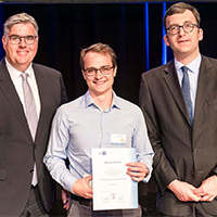 Drei Männer in Anzügen, einer hält eine Urkunde, stehen nebeneinander vor einem schwarzen Hintergrund.