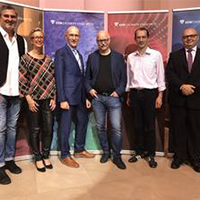 Sieben Personen stehen vor bunten Bannern in formeller Kleidung nebeneinander und lächeln in die Kamera.