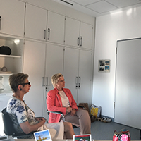 Zwei Frauen sitzen in einem hellen Büro mit weißen Schränken, einer trägt ein rosa Sakko, die andere ein weißes Kleid mit Blumenmuster.