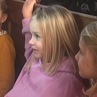 Ein kleines Mädchen in rosa Pullover sitzt neben anderen Kindern und schaut aufmerksam zur Seite.