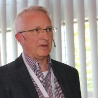 Ein Mann mit Brille hält einen Vortrag vor einem beschrifteten Banner, Hintergrund mit vertikalen Jalousien.