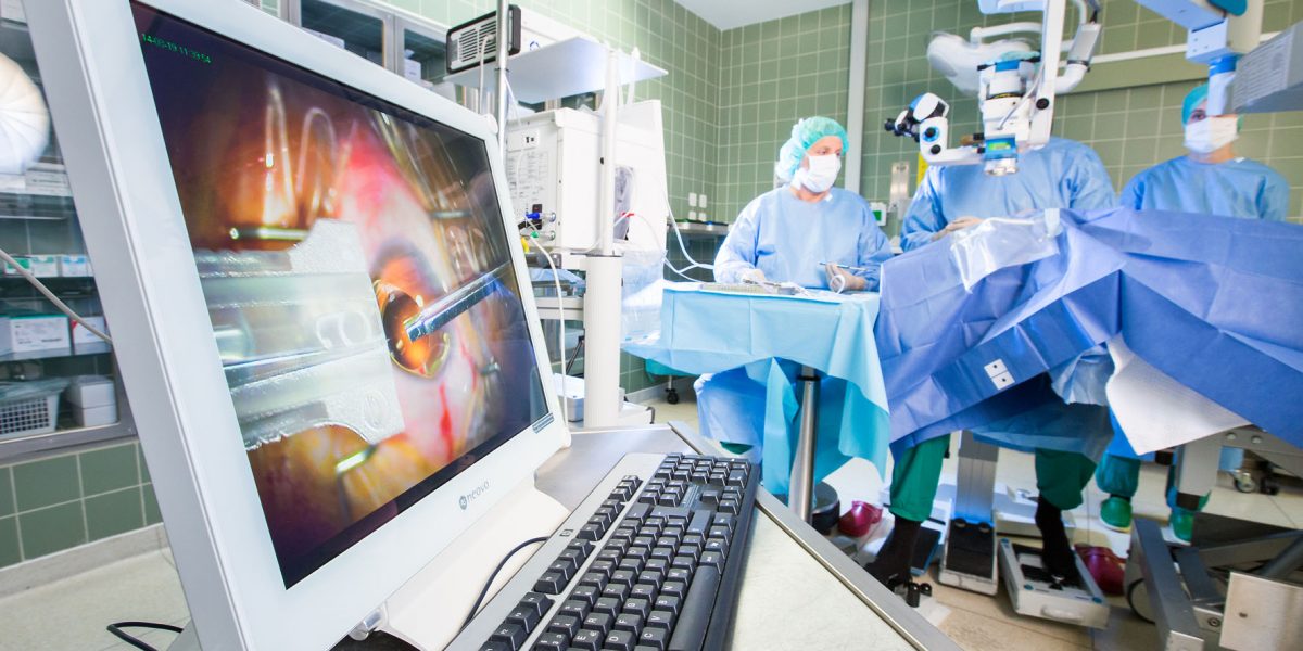 OP-Saal mit Monitor, der eine intraokulare Operation zeigt, während Ärzte in OP-Kleidung den Eingriff durchführen.