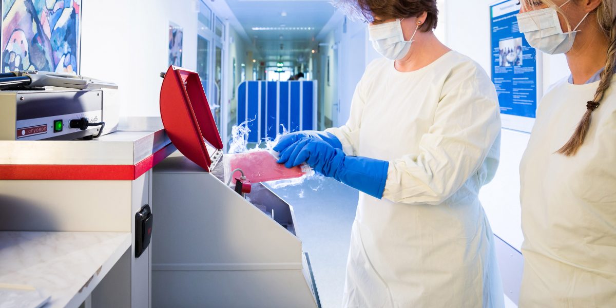 Zwei maskierte Frauen in Schutzkleidung arbeiten an einem Kryogerät in einem Laborflur.