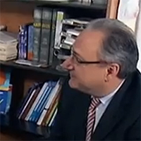 Mann im Anzug mit Brille sitzt in Büro vor Bücherregal, das mit verschiedenen Büchern und Akten gefüllt ist.