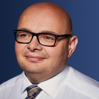 Ein Mann mit Brille im Anzug lächelt vor einem blauen Hintergrund.