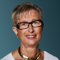 Porträt einer älteren Frau mit Kurzhaarfrisur, Brille und hellblauem Hintergrund. Sie trägt ein weißes Oberteil.