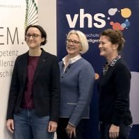 Drei Frauen stehen lächelnd vor Schildern der VHS und EM. im Innenraum, linke Frau im Blazer, mittlere Frau graumeliert.