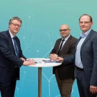 Drei Männer in Anzügen stehen an einem runden Tisch vor türkisfarbenem Hintergrund mit Liniengrafik.