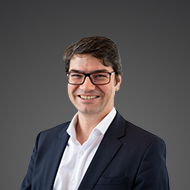 Ein lächelnder Mann mit Brille und dunklem Anzug vor grauem Hintergrund.