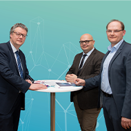 Drei Männer in Anzügen stehen an einem Tisch vor einem türkisfarbenen Hintergrund mit geometrischen Linien.