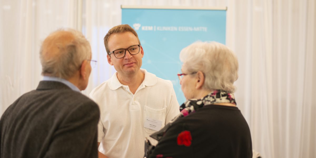 Mann im weißen Polohemd spricht mit zwei älteren Personen vor einem blauen Hintergrund mit Klinik-Logo.