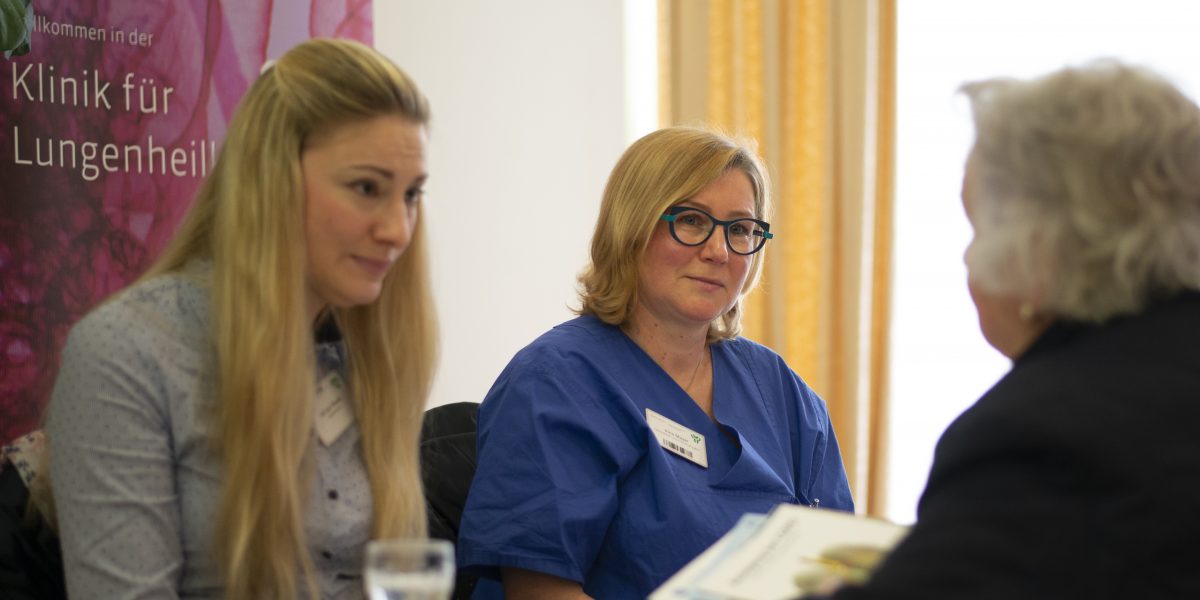 Drei Frauen in einem Gespräch, eine im blauen Kittel, vor einem Banner der Klinik für Lungenheilkunde.