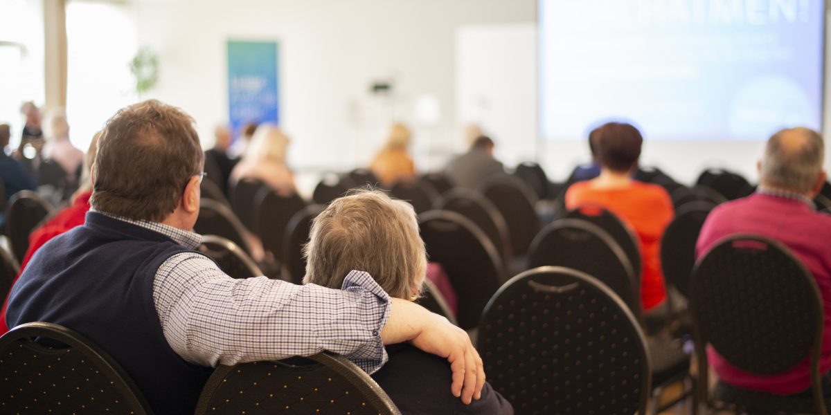 Personen in einem Konferenzraum, einige leere Stühle; ein Mann legt den Arm um eine ältere Person.