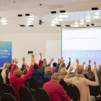 Leute in einem Seminarraum heben die Arme. Präsentation zu Luftschadstoffen und Gesundheit. Plakat: 