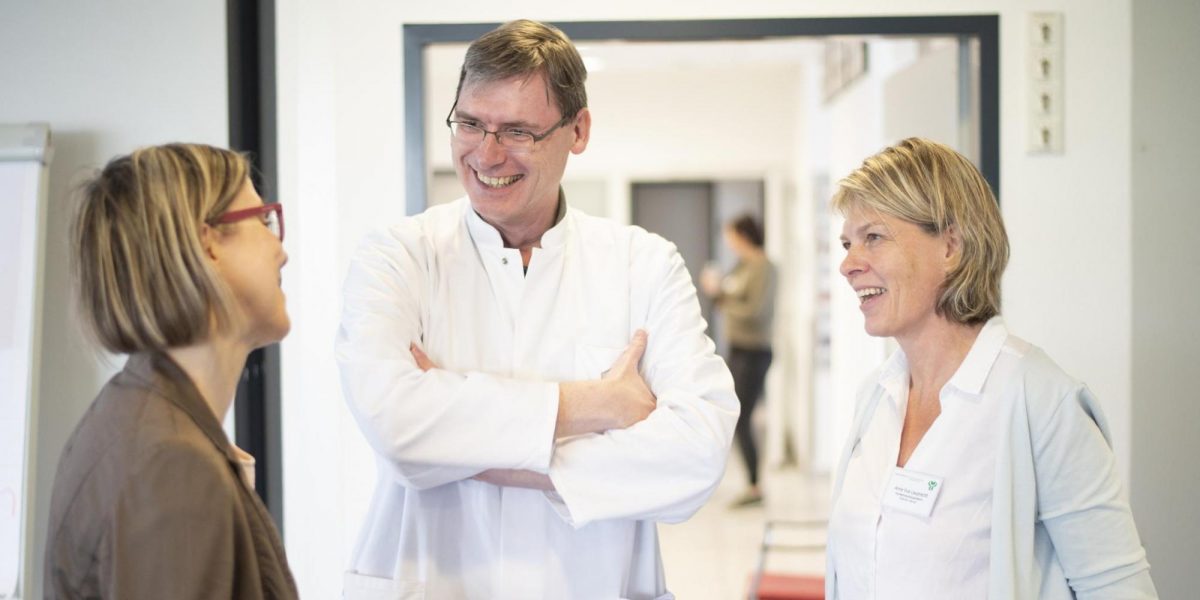 Drei Personen unterhalten sich freundlich in einem hellen Flur, eine davon trägt einen weißen Arztkittel.