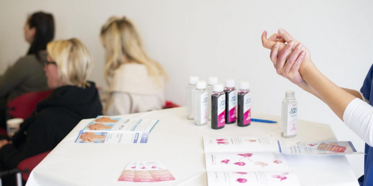 Person demonstriert Händedesinfektion an Tisch mit Desinfektionsmittel und Infomaterial, während andere im Hintergrund sitzen.