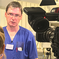 Mann in blauer Arztkleidung spricht vor einer Kamera.