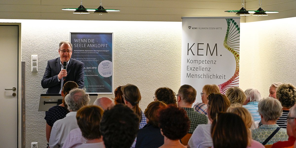 Ein Mann hält eine Rede vor Zuhörern, neben einem Poster mit der Aufschrift