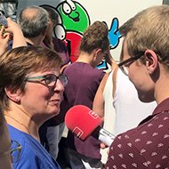 Eine Frau wird auf der Straße von einem Reporter mit Mikrofon interviewt; Hintergrund mit Comic-Graffiti.