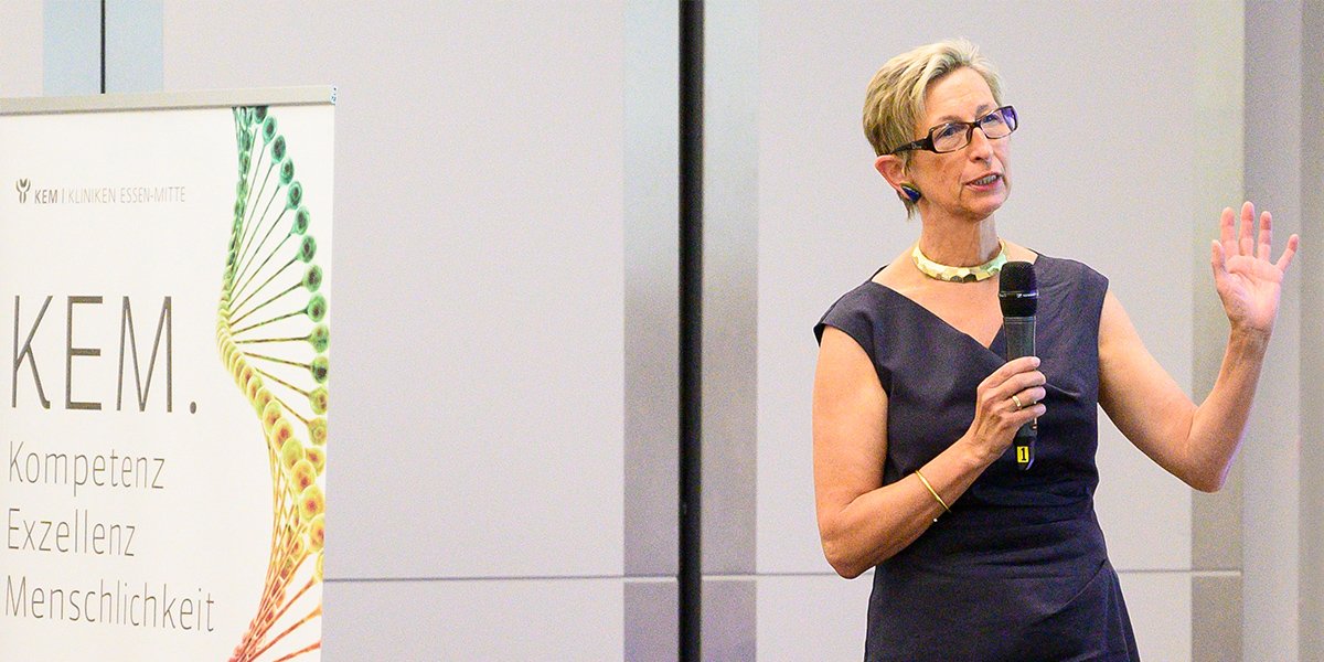 Frau hält Vortrag mit Mikrofon vor KEM-Banner. Text: