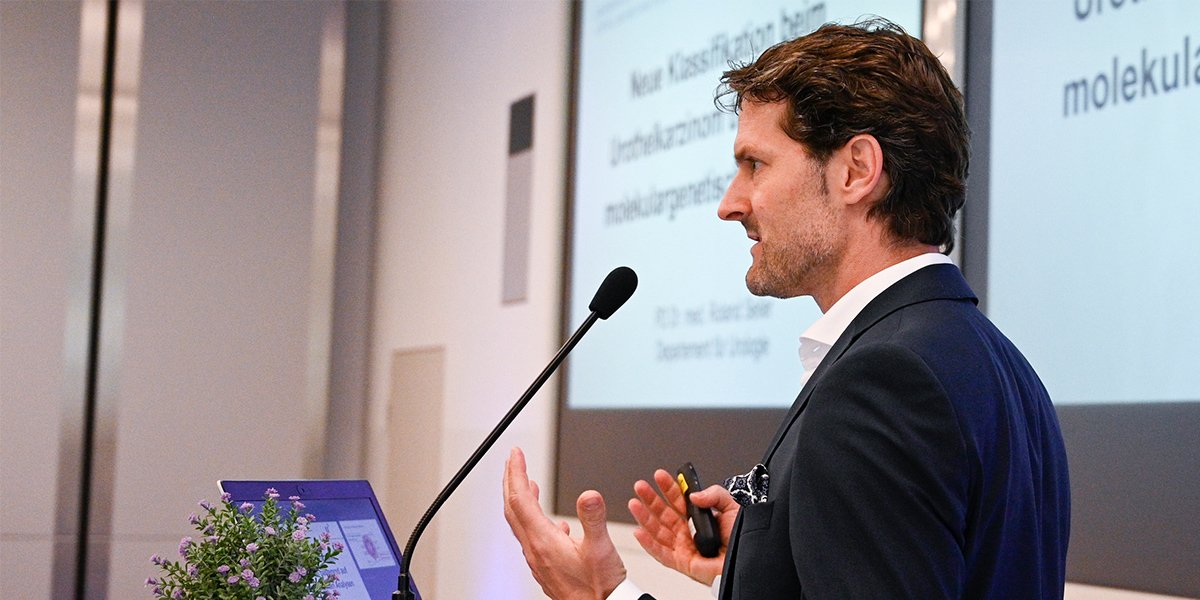 Mann hält einen Vortrag neben einem Mikrofon und Bildschirm mit Präsentationstext. Bildschirm zeigt unleserliche Schrift.