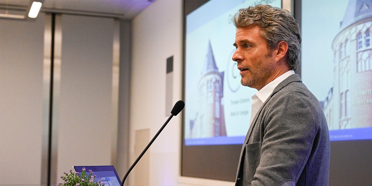 Mann hält eine Präsentation in einem Konferenzraum, Leinwand im Hintergrund, Laptop auf einem Tisch vor ihm.