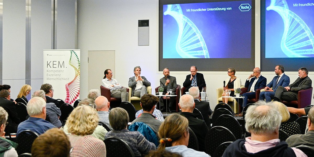 Podiumsdiskussion mit acht Personen auf einer Bühne, Publikum im Vordergrund, DNA-Bild auf der Leinwand.