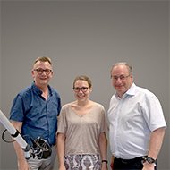 Drei Personen stehen lächelnd nebeneinander vor grauem Hintergrund. Links ist ein Gerät teilweise sichtbar.