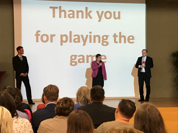 Drei Personen auf einer Bühne, zweisprachig mit Mikrofonen, vor einer Folie mit der Aufschrift: 