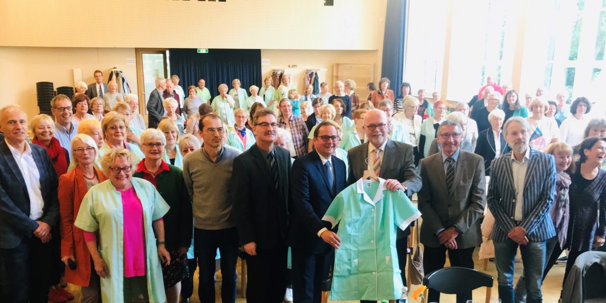 Gruppe von Personen in einem Raum, vorne sind vier Männer und eine Pflegekraft, die einen Kittel präsentieren.