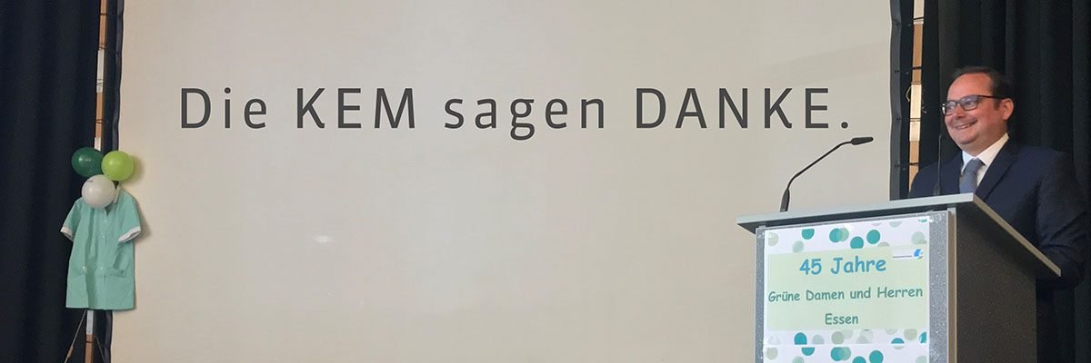 Ein Mann steht lächelnd am Rednerpult neben einer Leinwand mit der Aufschrift „Die KEM sagen DANKE“.