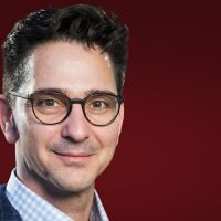 Mann mit Brille und Anzug vor dunkelrotem Hintergrund.