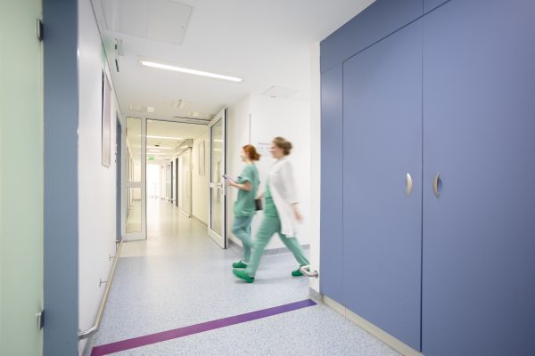 Zwei Personen in grüner Arbeitskleidung gehen schnell durch einen hellen Krankenhausflur mit blauem Schrank.