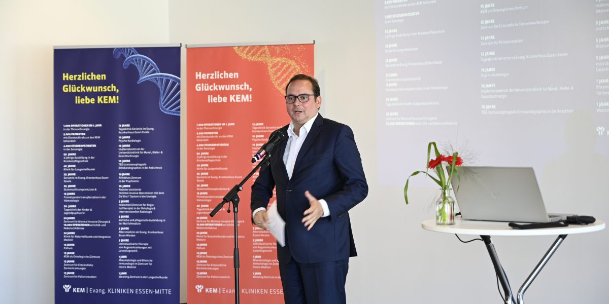 Ein Mann in Anzug spricht bei einer KEM-Veranstaltung vor zwei bunten Bannern, daneben ein Tisch mit Laptop und Blumen.