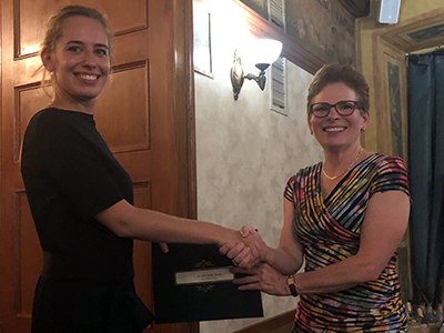 Zwei Frauen schütteln sich die Hand. Eine hält eine dunkle Mappe. Sie stehen vor einer hölzernen Tür in einem Zimmer.