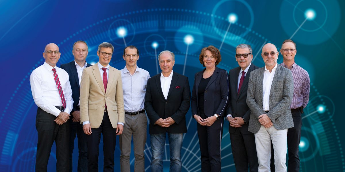 Neun Personen stehen nebeneinander vor einem blauen, technischen Hintergrund. Sie tragen formelle Kleidung.