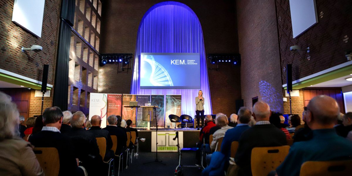 Eine Frau spricht auf einer Bühne in einer Halle vor einem sitzenden Publikum, im Hintergrund ein großer Bildschirm mit KEM-Logo.