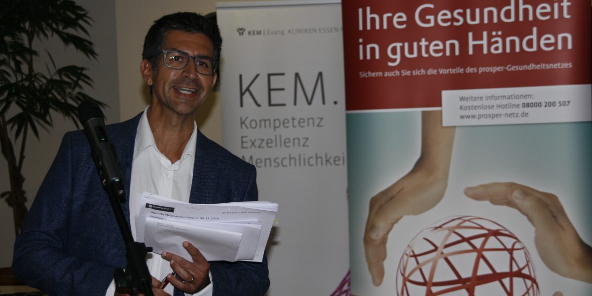 Mann hält Papiere vor einem Mikrofon; im Hintergrund ein KEM-Plakat und ein Banner mit 