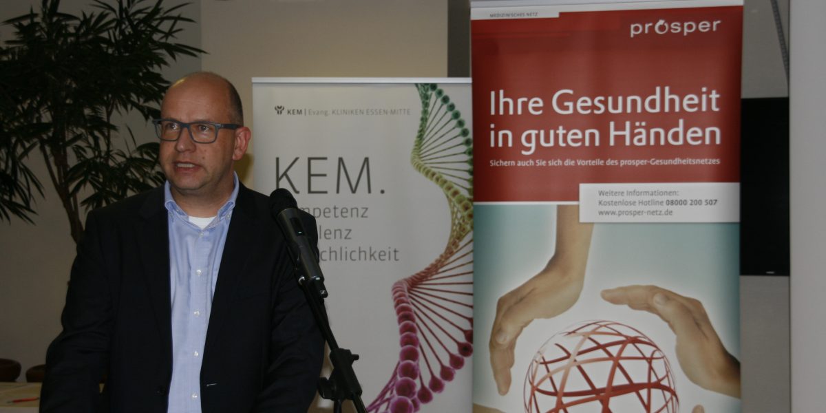 Ein Mann spricht bei einer Veranstaltung, im Hintergrund Banner von KEM und prosper-Gesundheitsnetzwerk.