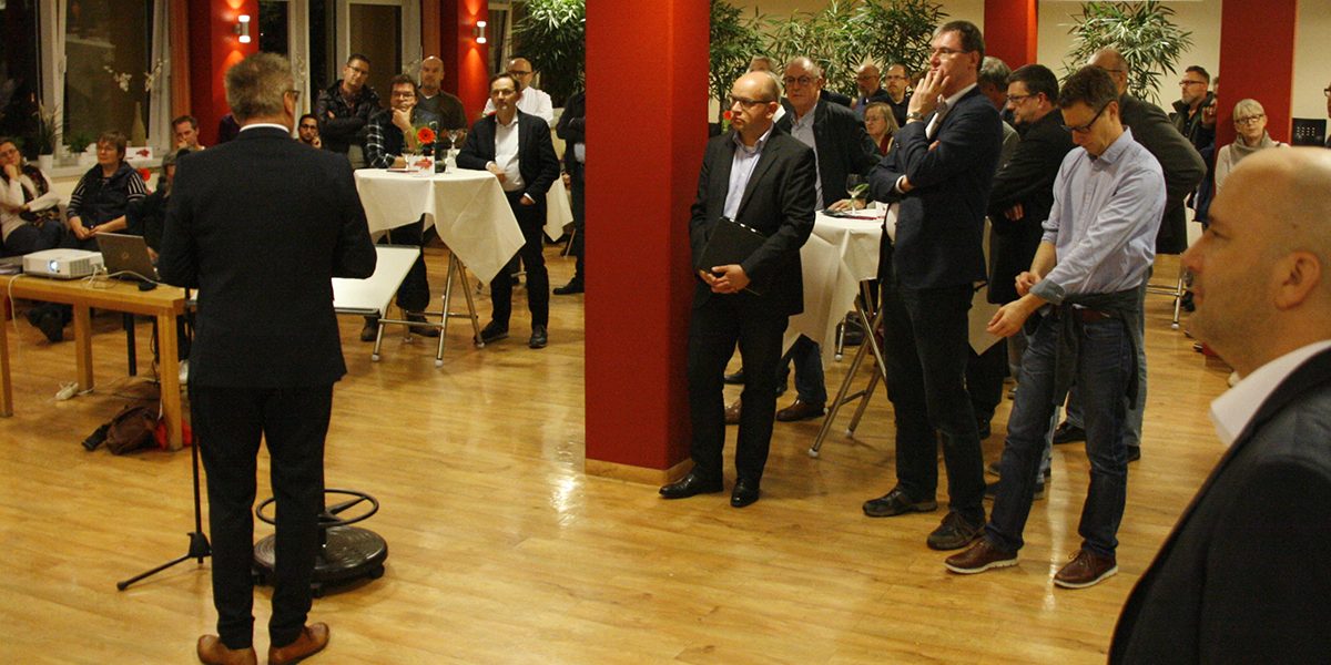 Person hält Rede vor Publikum in einem Raum mit roten Säulen und Stehtischen. Zuhörer stehen und sitzen aufmerksam.