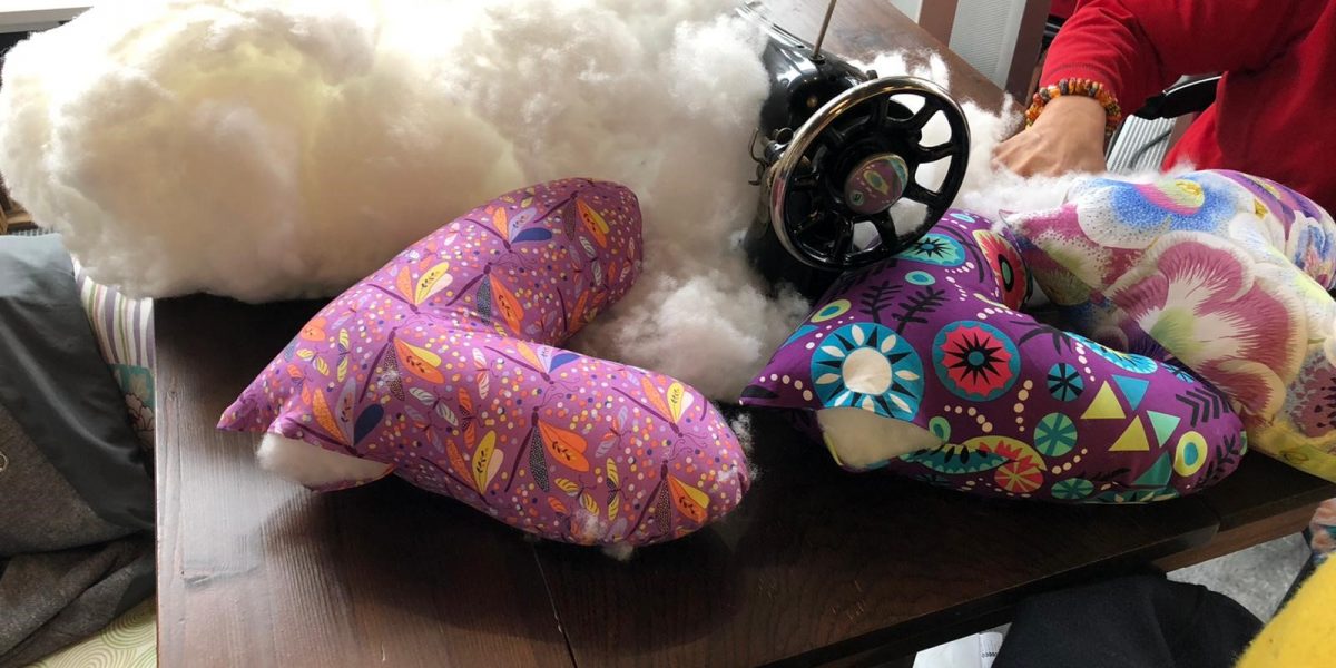 Nähmaschine mit bunten, mit Füllmaterial gefüllten Kissen auf einem Tisch. Hände einer Person daneben.