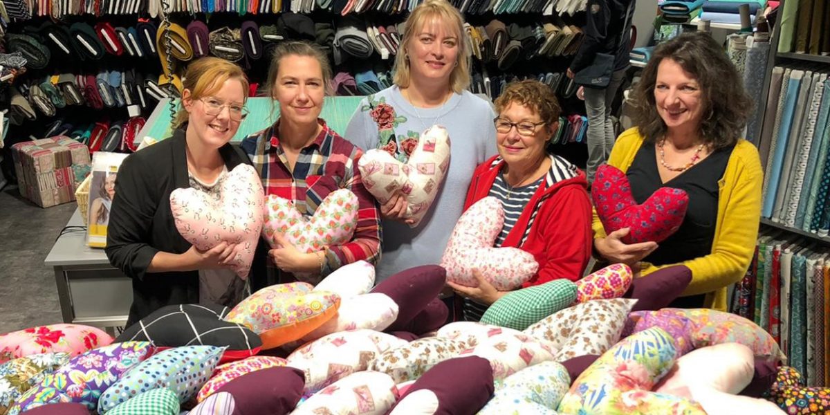 Fünf Personen halten herzförmige Kissen in einem Stoffgeschäft, umgeben von bunten Stoffballen.