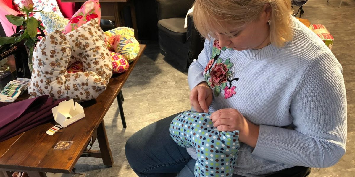 Frau näht an einem bunten Herzpolster, umgeben von Stoffmustern und Kissen in einer kreativen Werkstattumgebung.