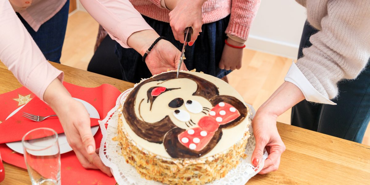Personen schneiden einen Kuchen mit einer Zeichentrickfigur darauf an. Tisch mit rotem Tischtuch und Geschirr.