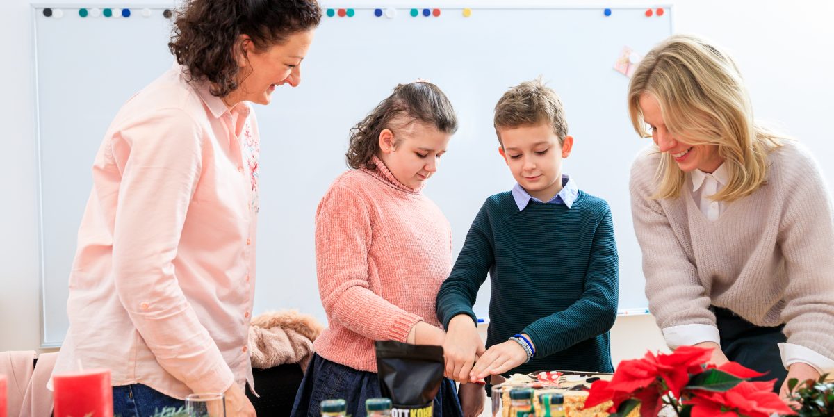 Vier Personen backen Kekse an einem festlich geschmückten Tisch mit weihnachtlichen Dekorationen im Vordergrund.