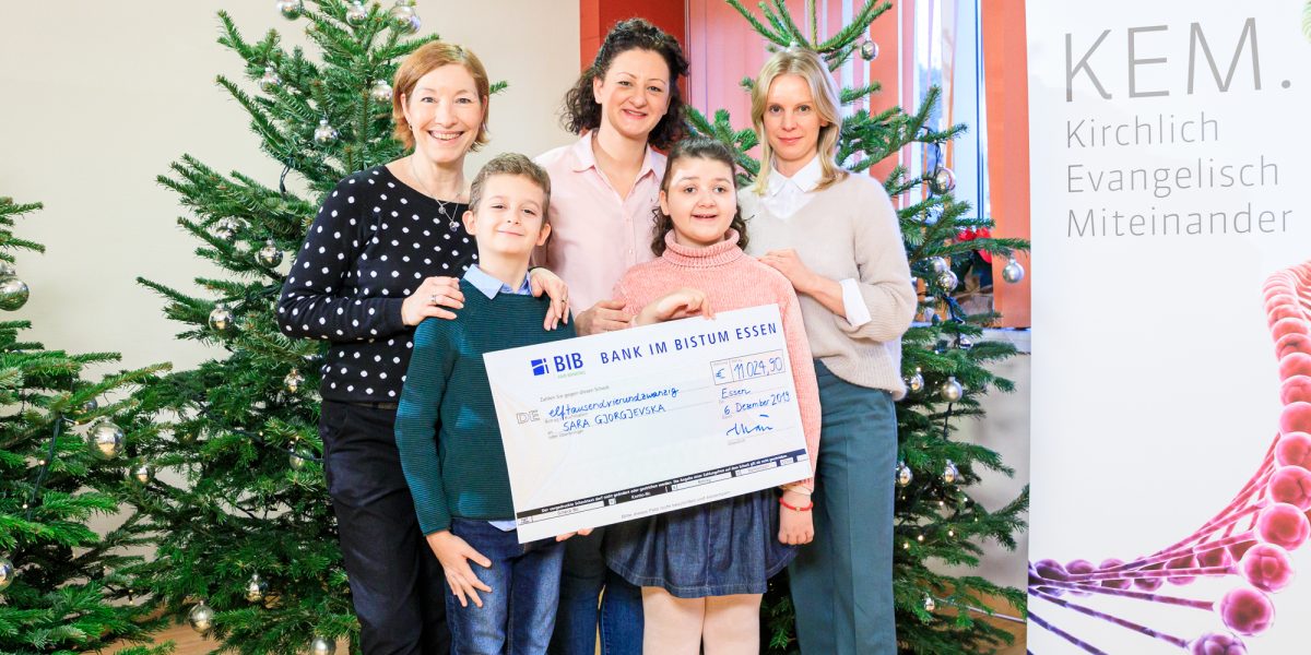 Fünf Personen mit übergroßem Scheck vor Weihnachtsbäumen und einem Banner der Kirchlich Evangelisch Miteinander.